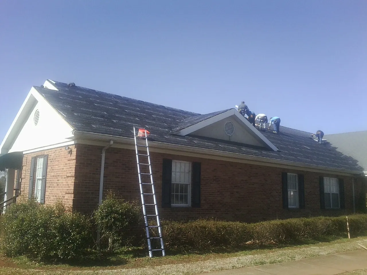 Crew tearing shingles off a brick ranch roof with a ladder set — Emergency Roof Repair work by our crew in Cazenovia