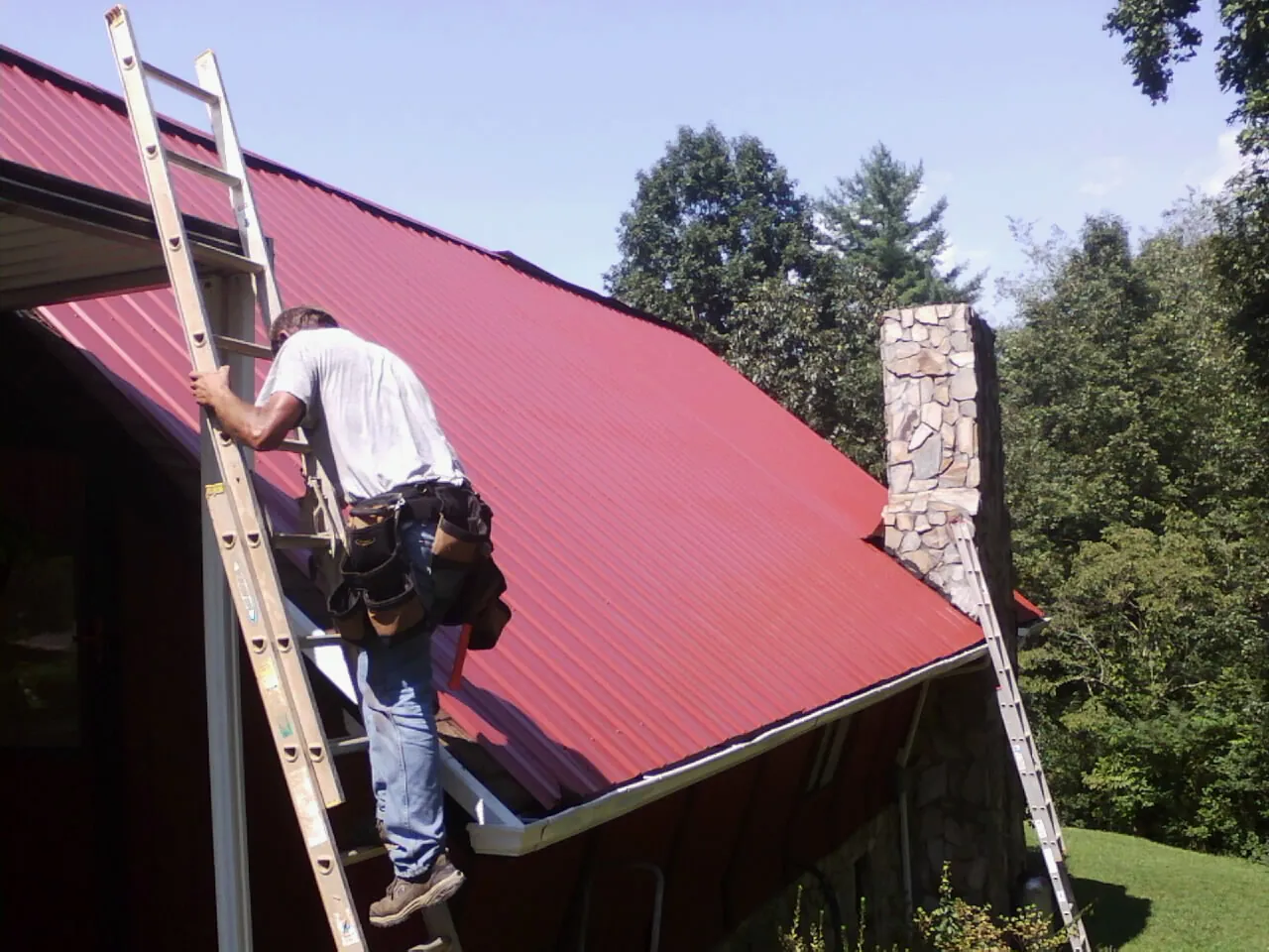 Roofer on a ladder beside a red metal roof and stone chimney — Soffit & Fascia Repair by Roofing Brothers in Cazenovia